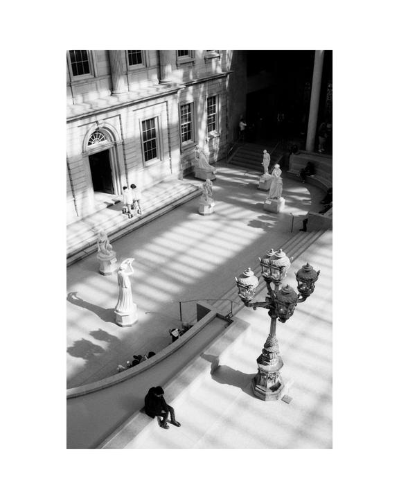 A high-angle view shows a large hall with statues, people, and a decorative lamppost, with strong shadows cast across the floor.