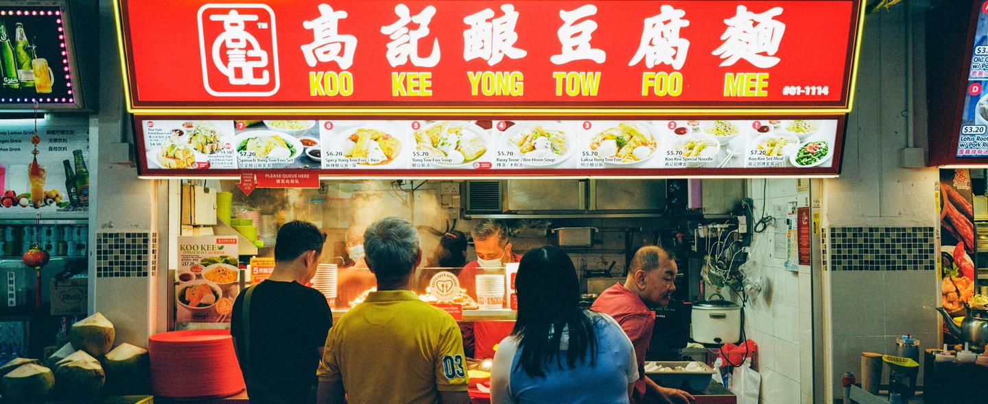 People stand in front of a food stall with a red sign and menu items displayed.