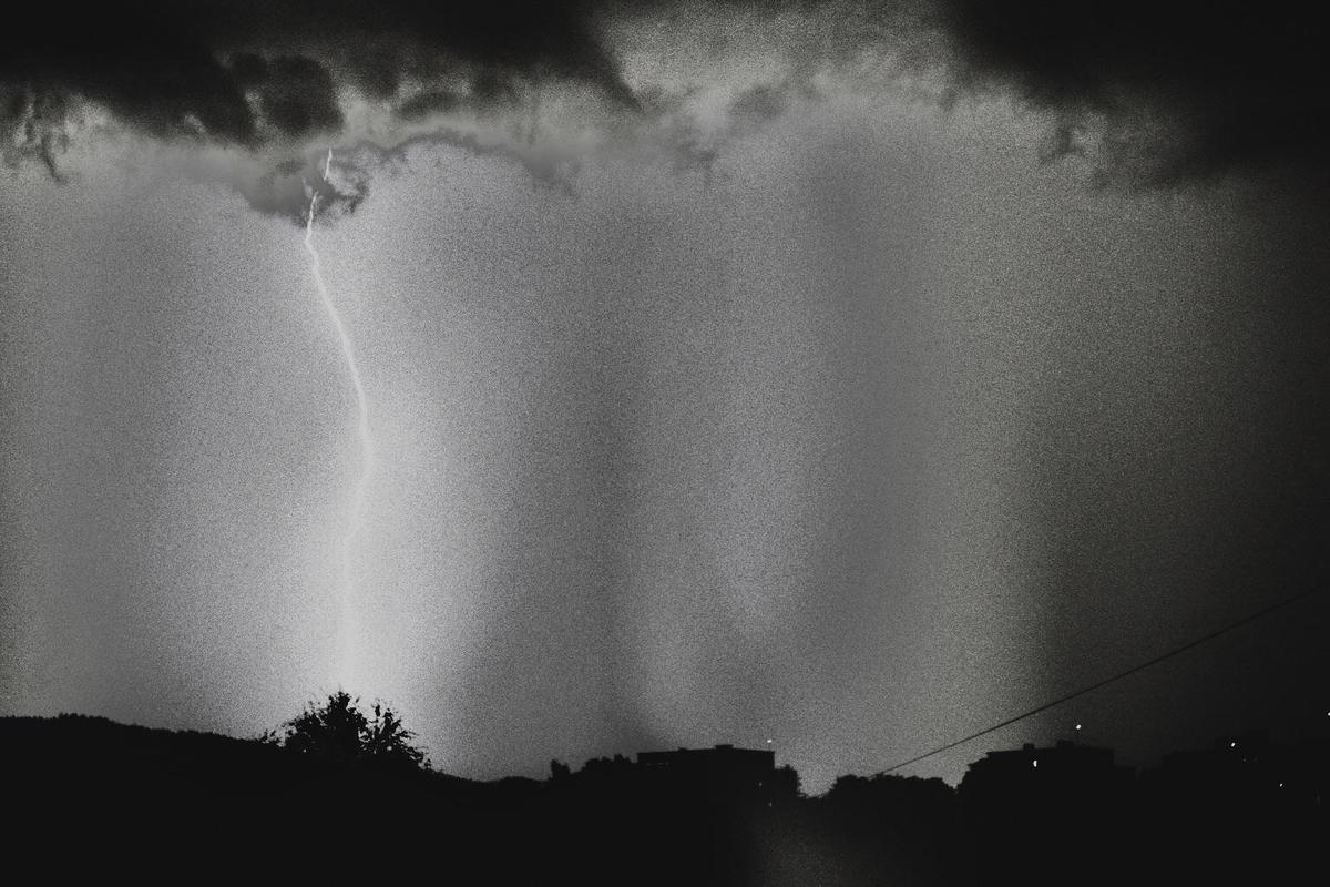 A bolt of lightning strikes from dark clouds above a silhouetted landscape with trees and buildings.
