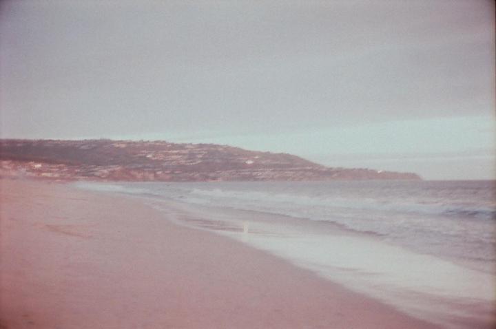 Waves crash on a sandy beach with a distant hillside covered in buildings under a hazy sky.