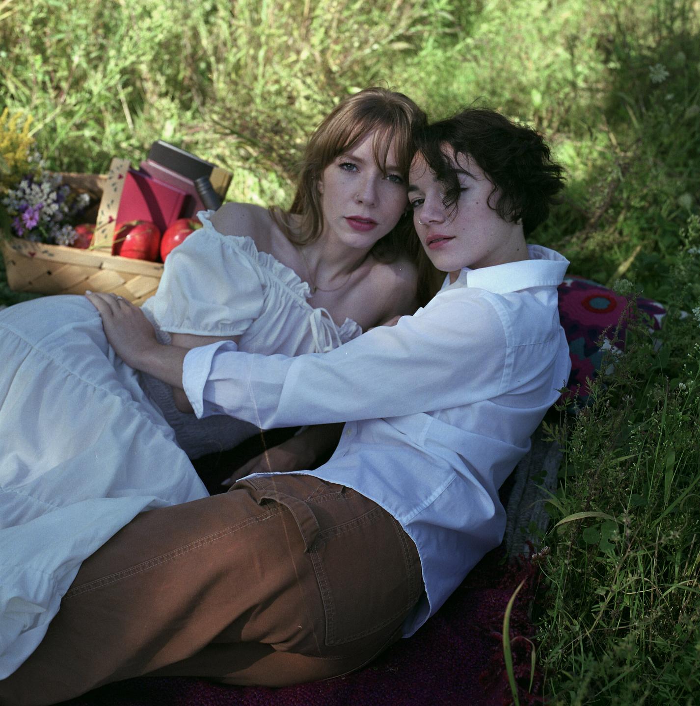 Two women are lying on a blanket in a grassy field with a picnic basket nearby.