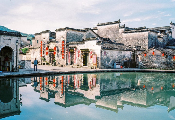 A man stands on the edge of a reflective pond in front of traditional Chinese buildings adorned with red lanterns.