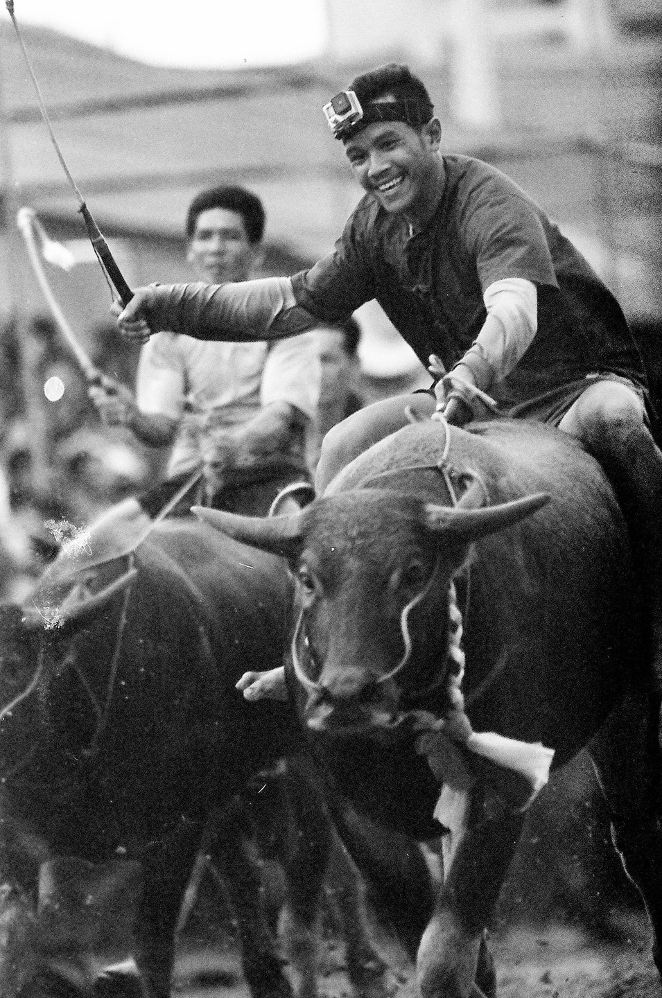 A man with a camera on his head smiles while riding a water buffalo during a race.