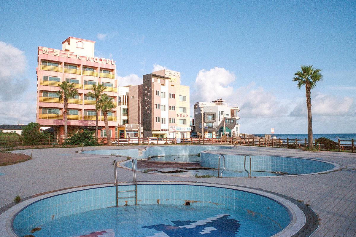Two empty swimming pools are in the foreground of a hotel and other buildings with the ocean in the background.