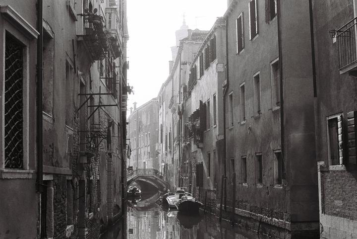 A narrow canal with boats and a bridge is surrounded by tall buildings in Venice.
