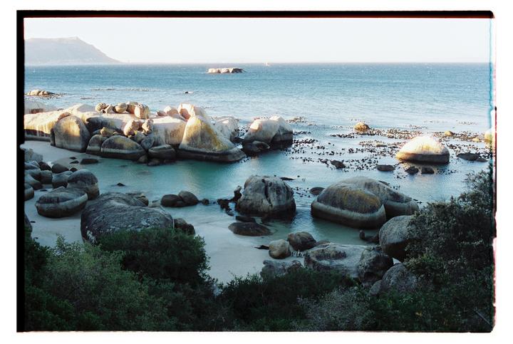 A beach with large boulders and calm water is visible, with a mountain in the distance.