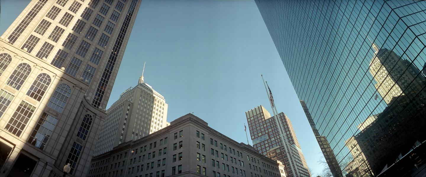 Looking up at several tall buildings, one with a reflective glass facade, under a clear sky.