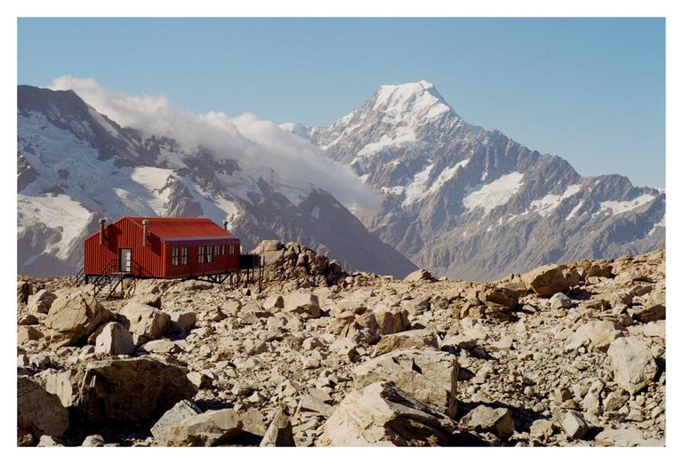 A red hut stands on a rocky, barren landscape with snow-capped mountains in the background under a clear sky.