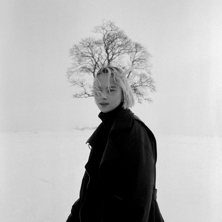 A woman with short hair and a dark coat stands in a snowy field with a bare tree in the background.