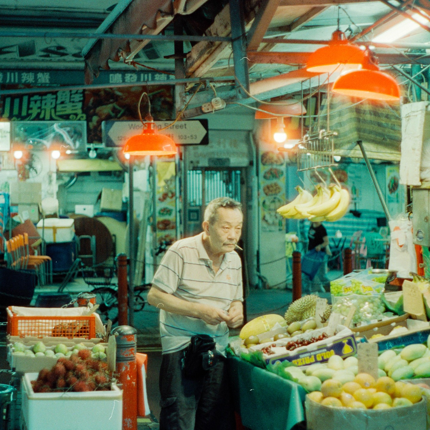 A man stands in a fruit stall at night, with bananas hanging above and various fruits displayed on tables.