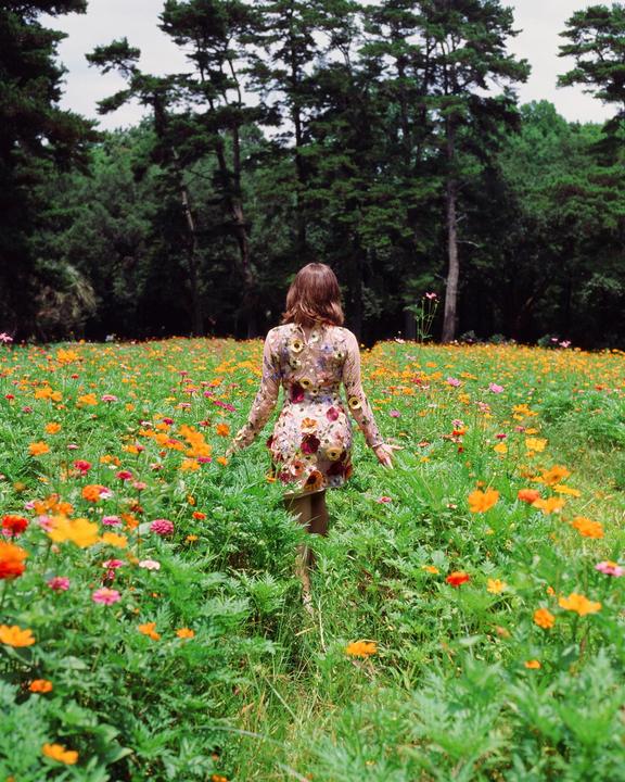 A woman in a floral dress walks through a field of wildflowers towards a forest.