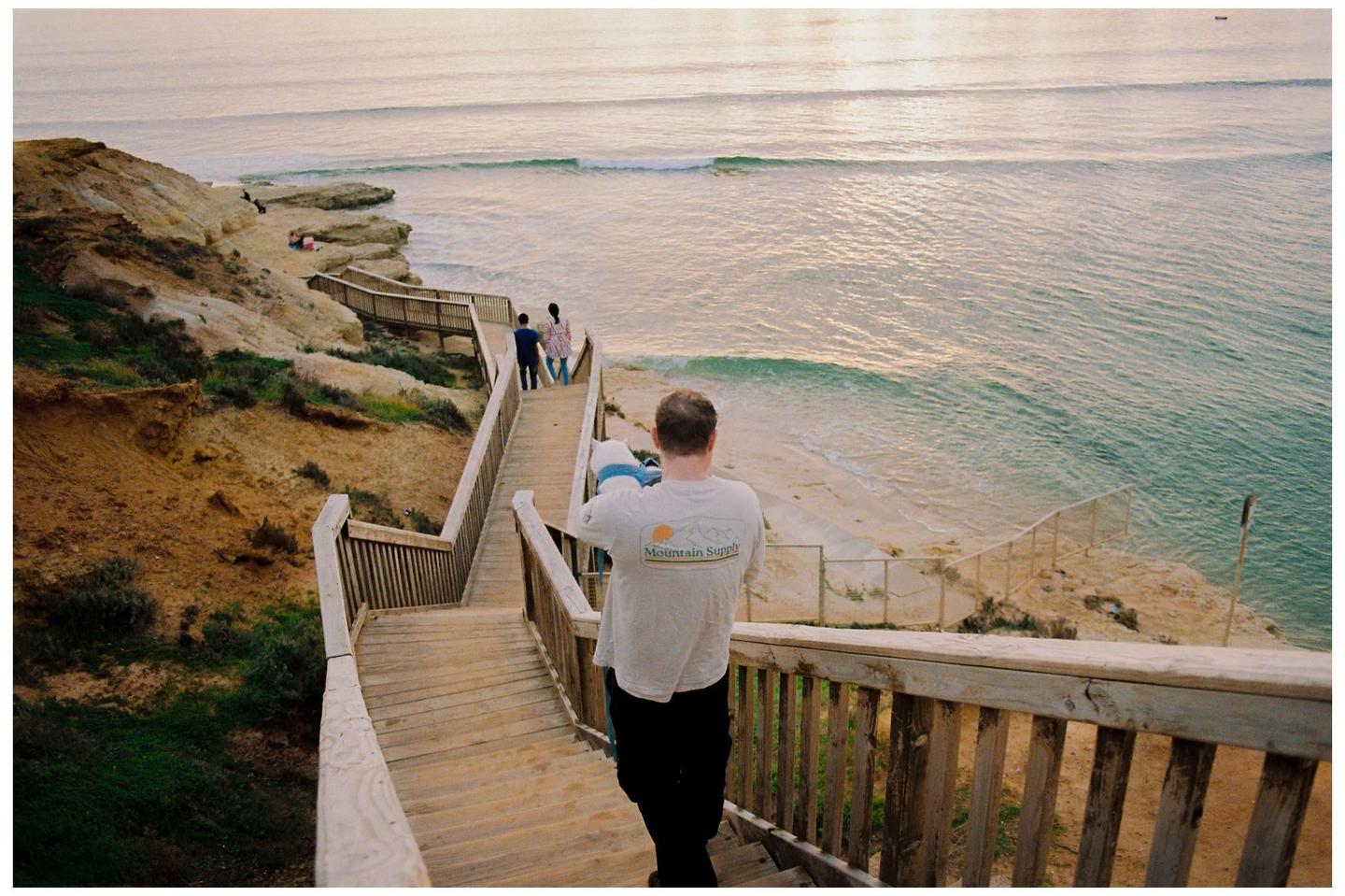 A man descends a long wooden boardwalk towards a beach with two people walking ahead of him.