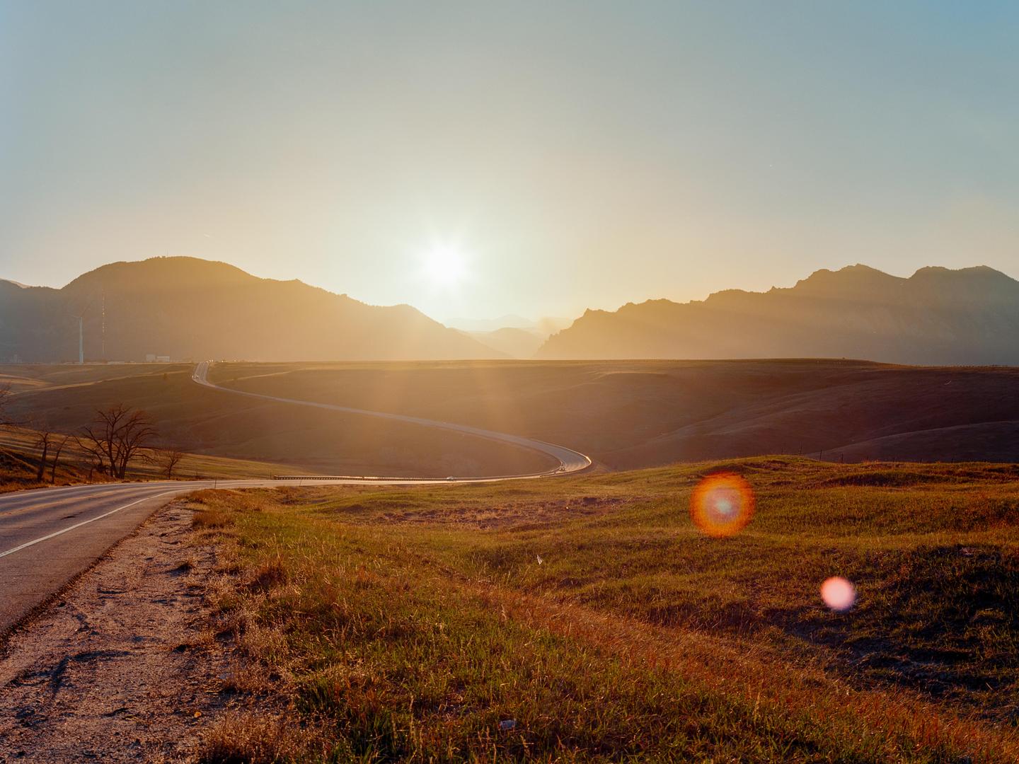 A winding road leads through a grassy landscape towards mountains under a bright sunset.