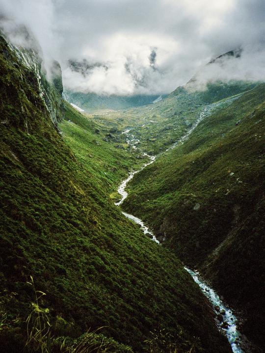 A river flows through a valley surrounded by mountains under a cloudy sky.