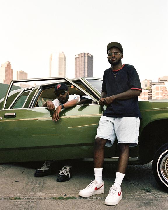 Two men pose with a green car on a street with a city skyline in the background.