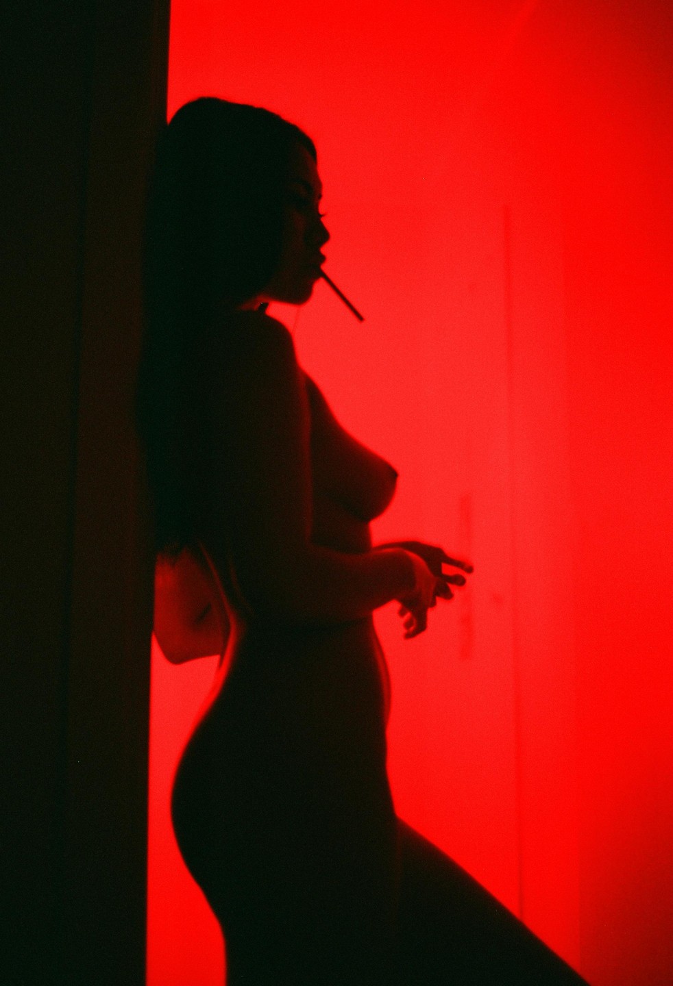A woman in silhouette with a cigarette in her mouth stands against a red wall.