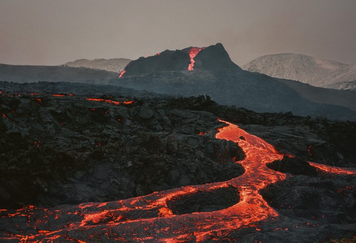 Molten lava flows from a volcanic mountain and across a dark, rocky landscape.