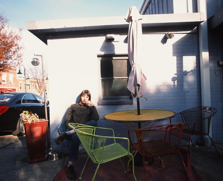 A woman sits in a green metal chair next to a table with an umbrella on a patio.