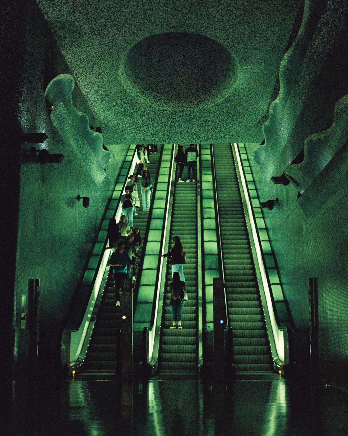 People ride up three escalators in a subway station with green lighting and a textured ceiling.