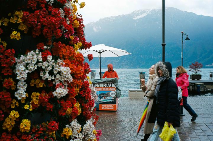 A person in an orange jacket stands by a "rent boat" sign next to a body of water with mountains in the background, while three people walk past a wall of colorful flowers in the foreground.