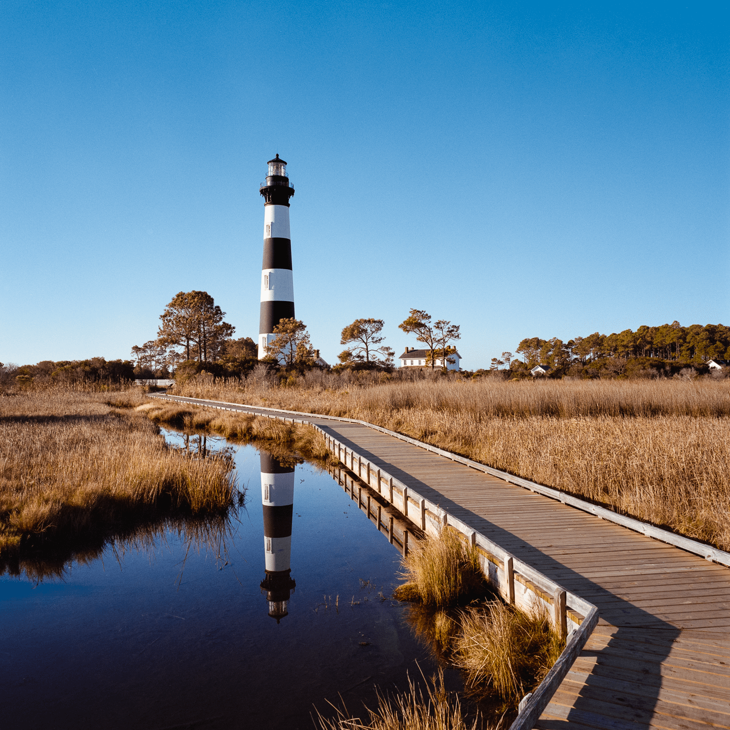 A black and white striped lighthouse stands tall behind a wooden boardwalk that stretches over a marsh with its reflection visible in the water.