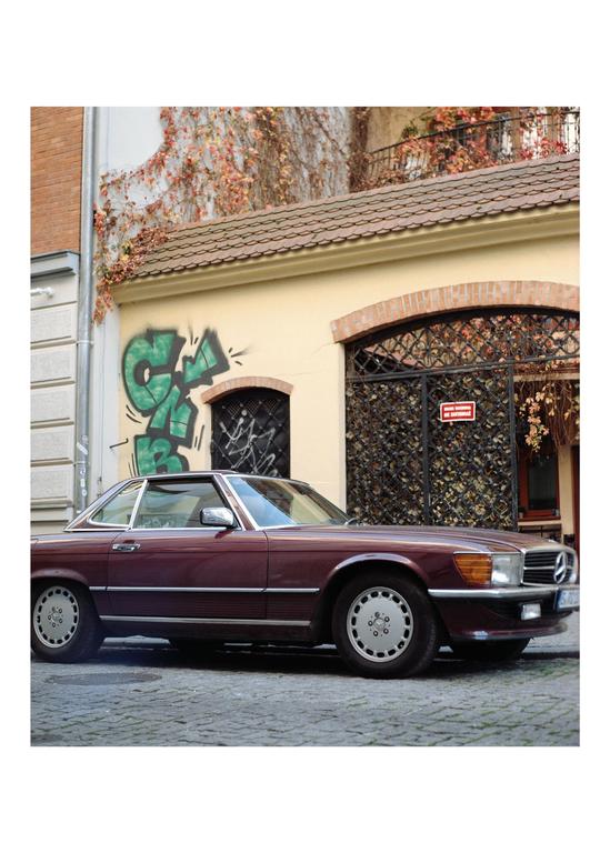 A maroon car is parked on a cobblestone street in front of a building with ivy and graffiti.