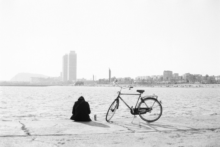 A person sits on a concrete pier next to a bicycle, looking out at the water and a distant city skyline.