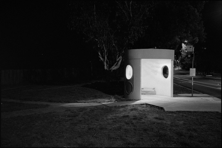A bus stop with a curved wall and two circular windows is illuminated at night next to a road and trees.