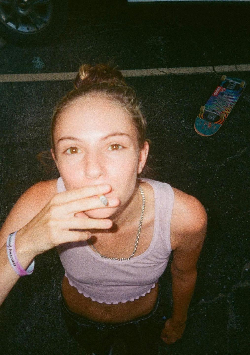 A young woman with her hair in a bun looks up at the camera while holding a cigarette to her mouth, with a skateboard visible on the ground beside her.