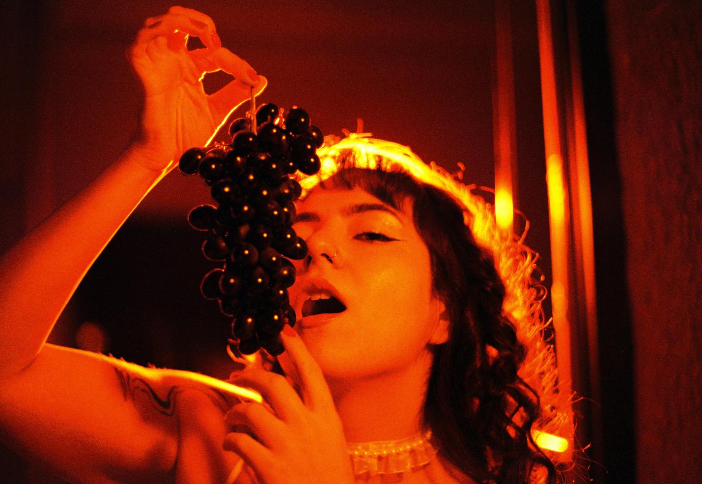 A woman holds a bunch of grapes to her mouth, illuminated by orange light.