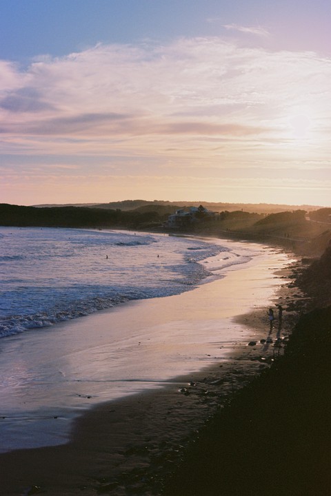A wide sandy beach curves along the coast with gentle waves rolling in under a pastel sunset sky.