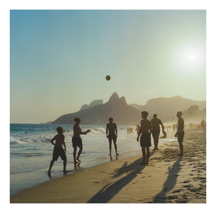 A group of men play volleyball on a beach at sunset with mountains in the background.