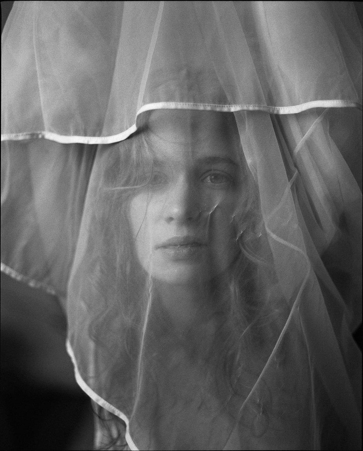 A woman with wavy hair looks directly forward from behind a sheer veil.