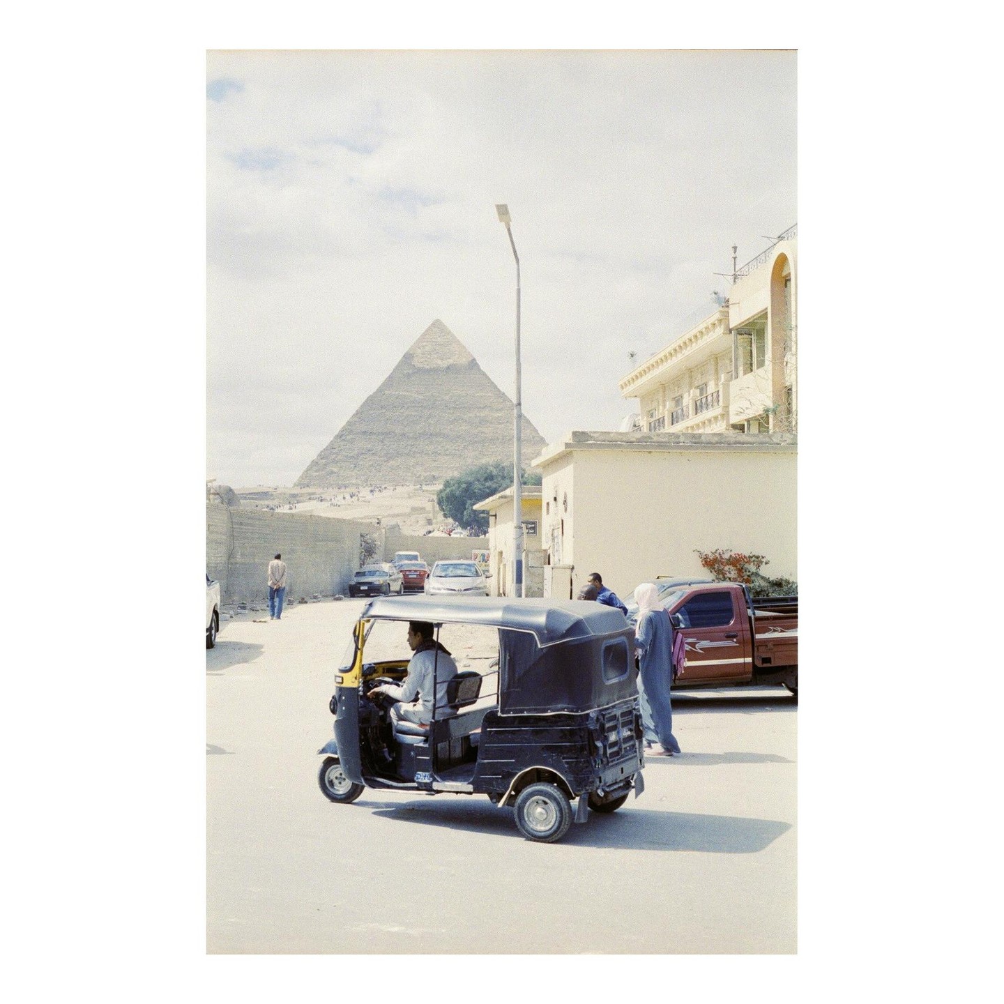 A man drives a tuk-tuk on a street with a pyramid in the background.