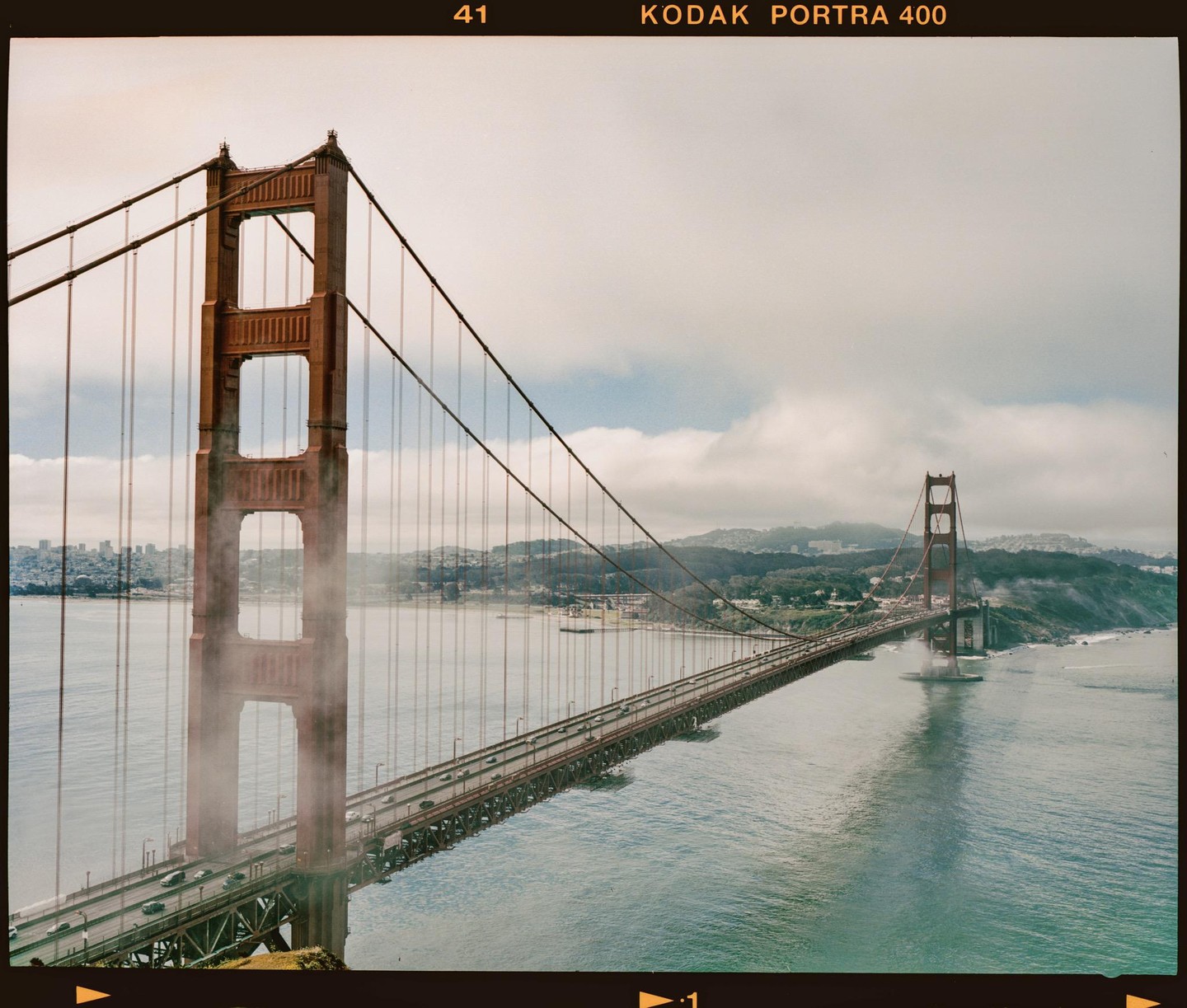 The Golden Gate Bridge is partially obscured by fog, with the San Francisco skyline visible in the distance under a cloudy sky.