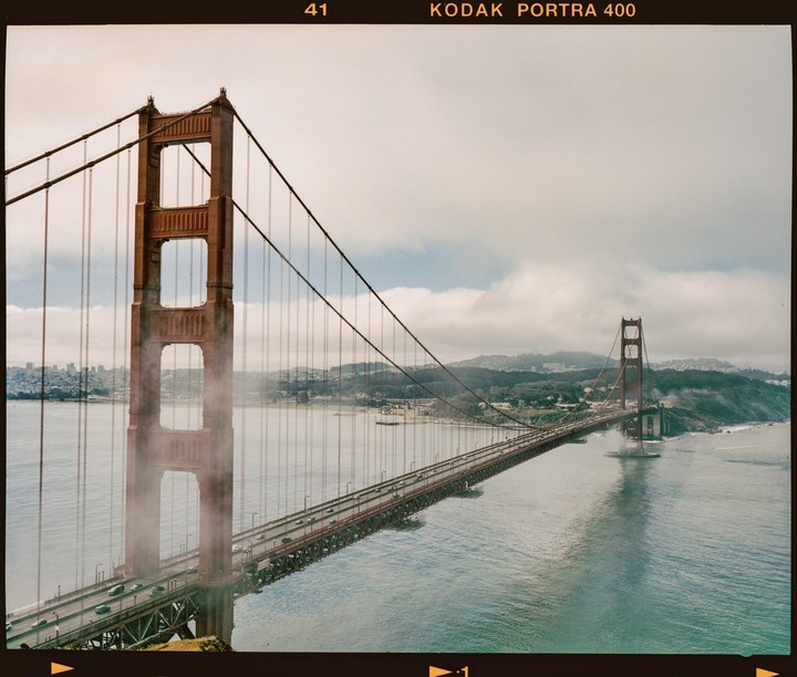 The Golden Gate Bridge is partially obscured by fog, with the San Francisco skyline visible in the distance under a cloudy sky.