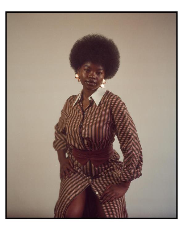 A woman with an afro wears a striped dress with a wide belt and large earrings.