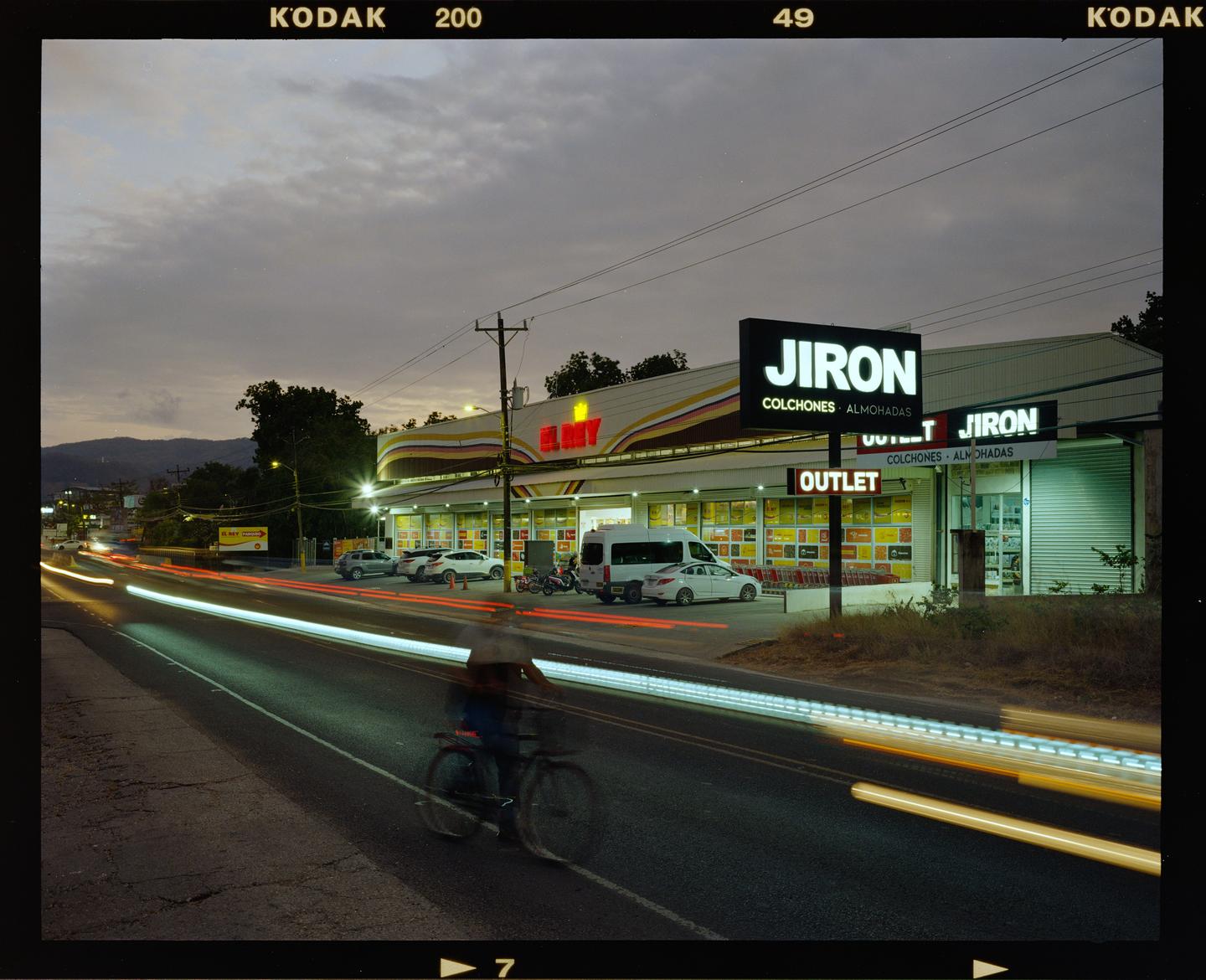 A cyclist rides past a brightly lit store with light trails from passing vehicles on the road at dusk.