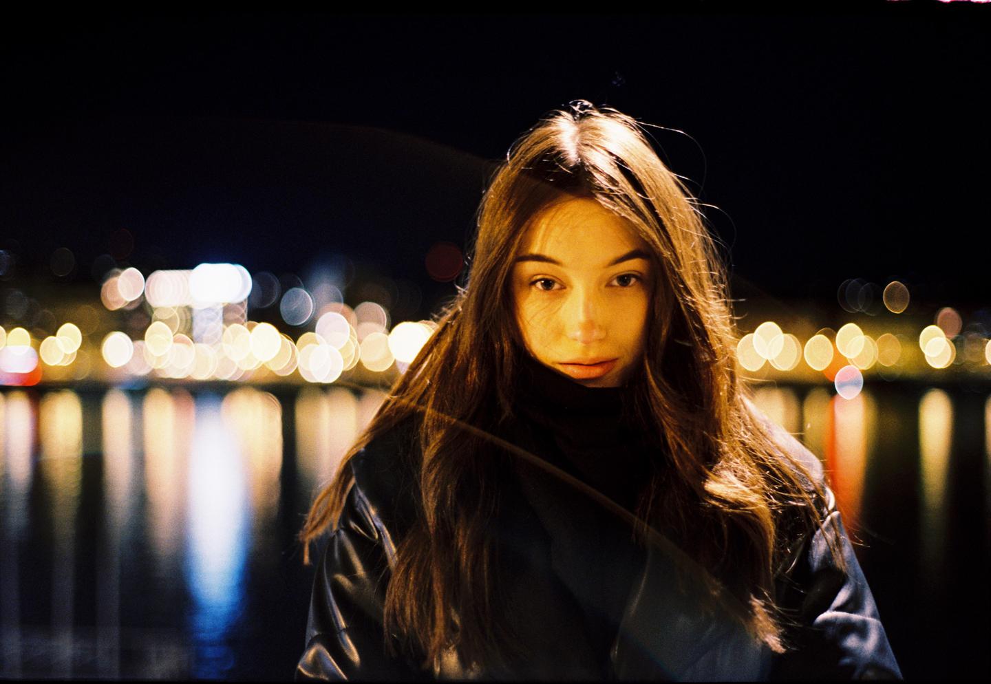 A woman with long hair looks directly at the viewer, illuminated by warm light against a dark background with blurred city lights reflecting on water.