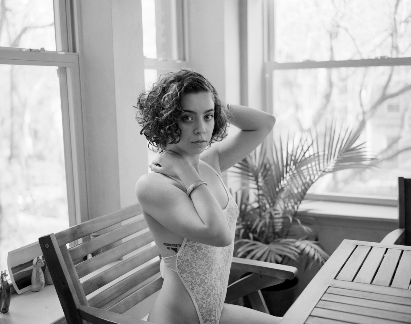 A woman with curly hair sits in a chair next to a window, wearing a lace bodysuit.