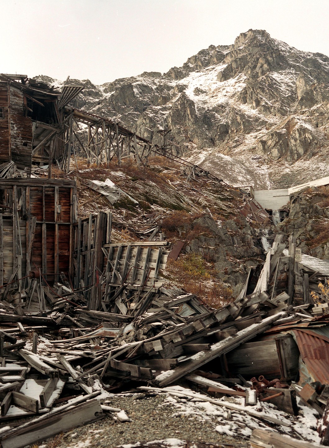 A ruined wooden structure stands on a snowy mountain slope with a waterfall and a snow-capped mountain in the background.