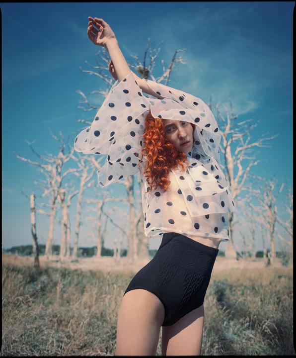 A woman with red hair and a polka dot top stands in a field with bare trees under a blue sky.