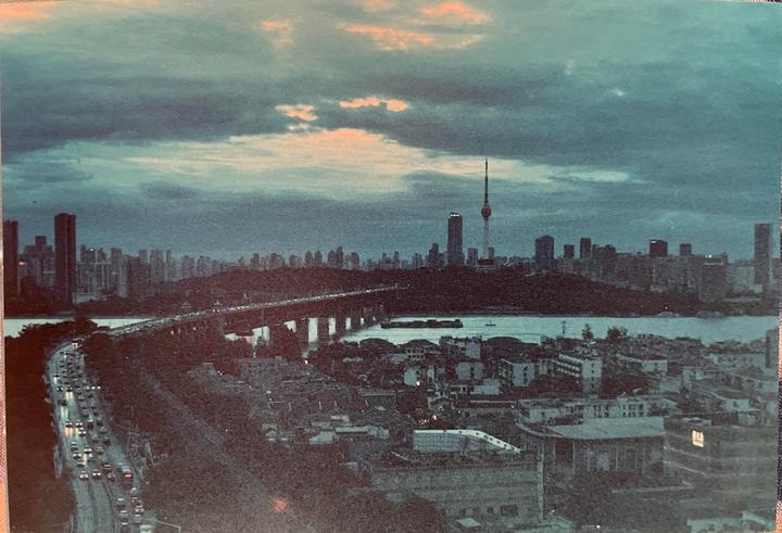 A city skyline with a river and a bridge is visible under a cloudy sky.