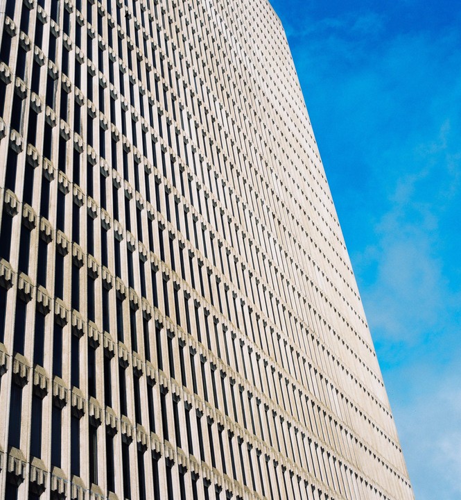 A tall building with many windows is shown from a low angle against a bright blue sky.