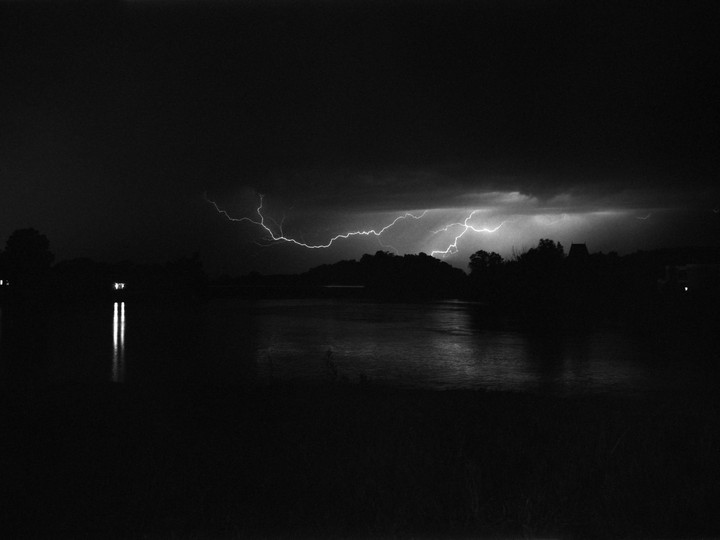 Lightning strikes across a dark sky above a body of water and silhouetted trees.