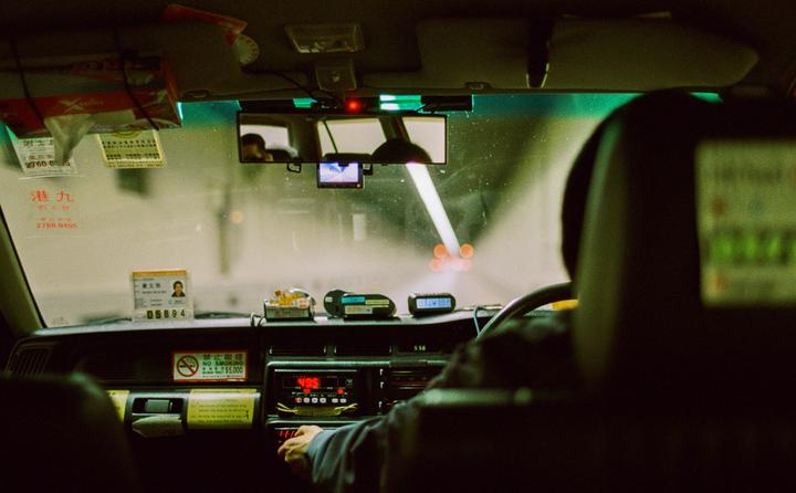 The interior of a taxi is visible from the back seat, with the driver's headrest and dashboard in the foreground and a tunnel visible through the windshield.