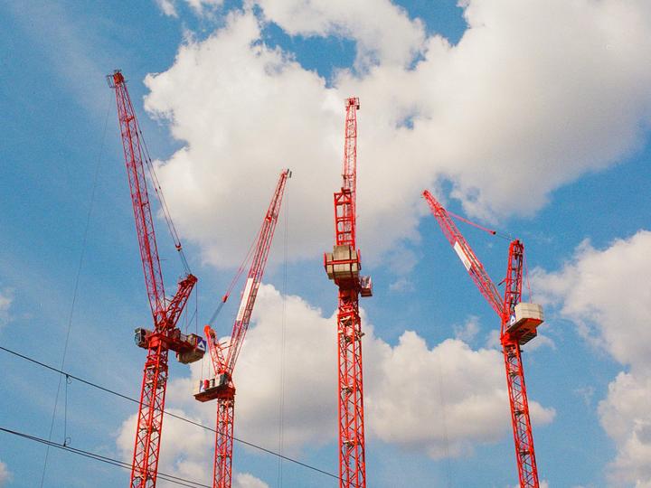 Four red construction cranes stand against a blue sky with white clouds.