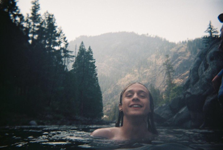 A smiling man with long hair is swimming in a body of water surrounded by a forest and mountains.