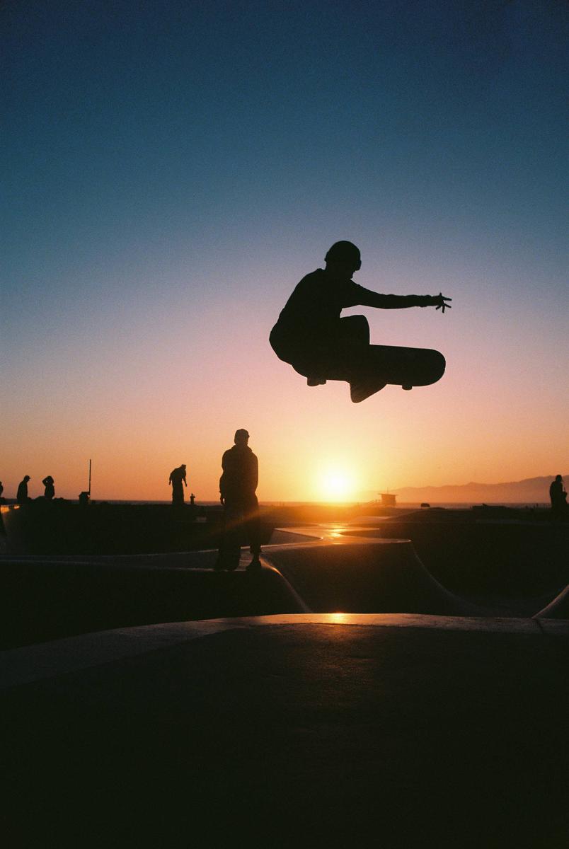 A skateboarder is silhouetted mid-air against a sunset sky at a skate park.