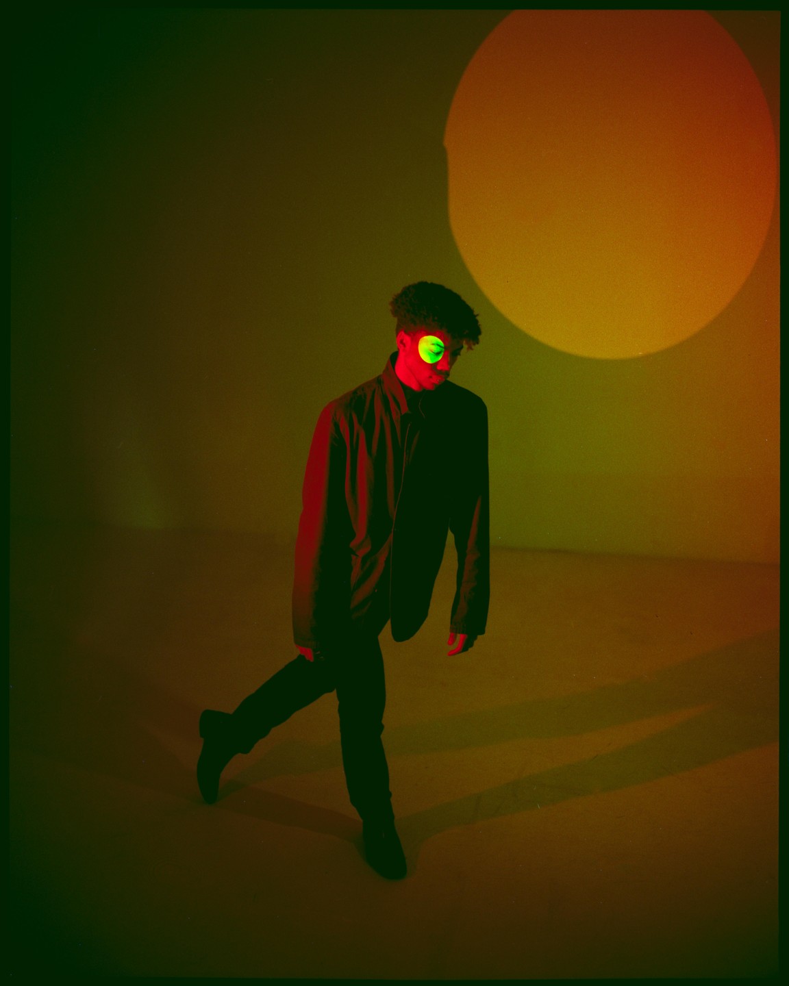 A man with a green circle over his eye stands in a room with red and yellow lighting.
